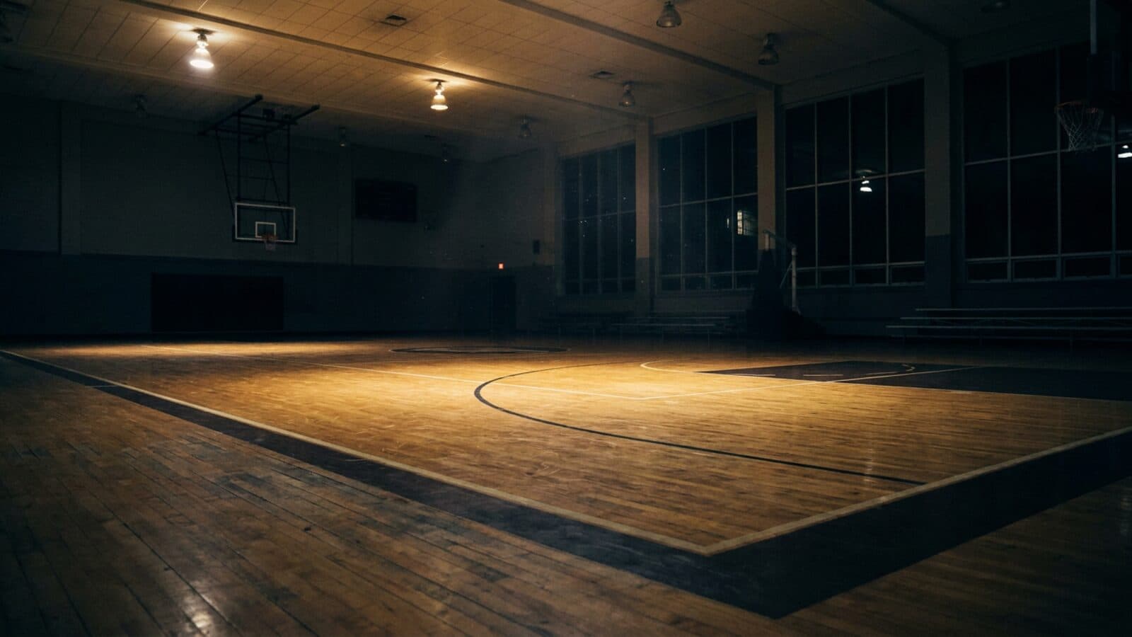 An indoor basketball court at night lit only by the overhead court lights, with the surrounding gym falling away into darkness