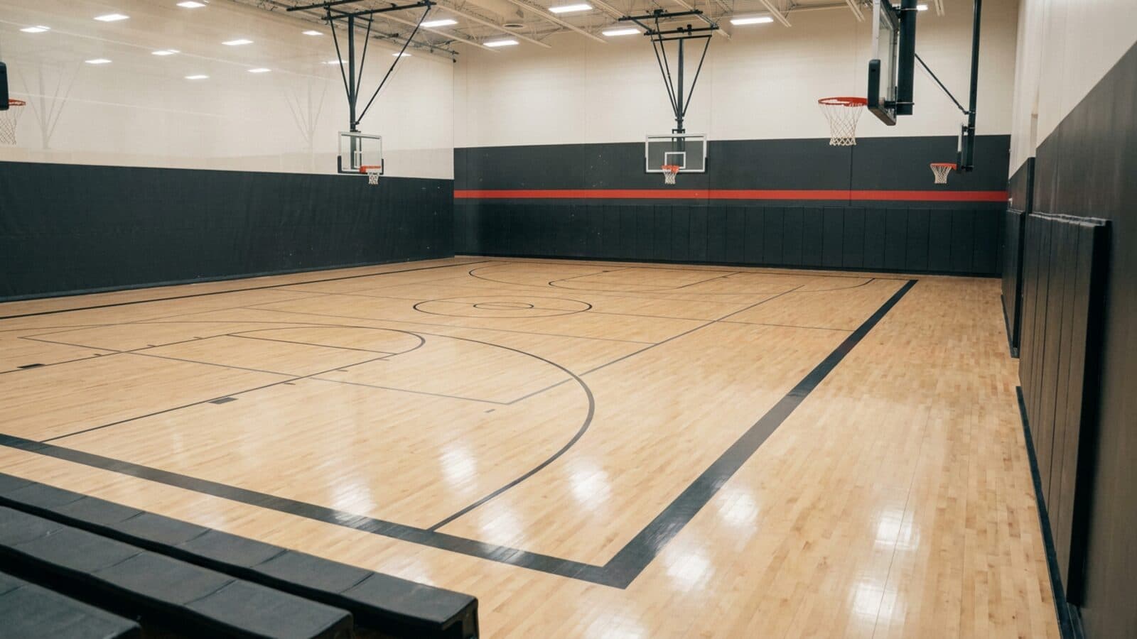 An empty indoor basketball gymnasium with a pale hardwood floor and two hoops receding down the hall under even overhead lighting