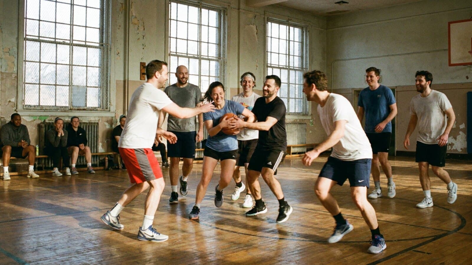 An indoor basketball court with a group mid-game on a winter afternoon, daylight low through high windows