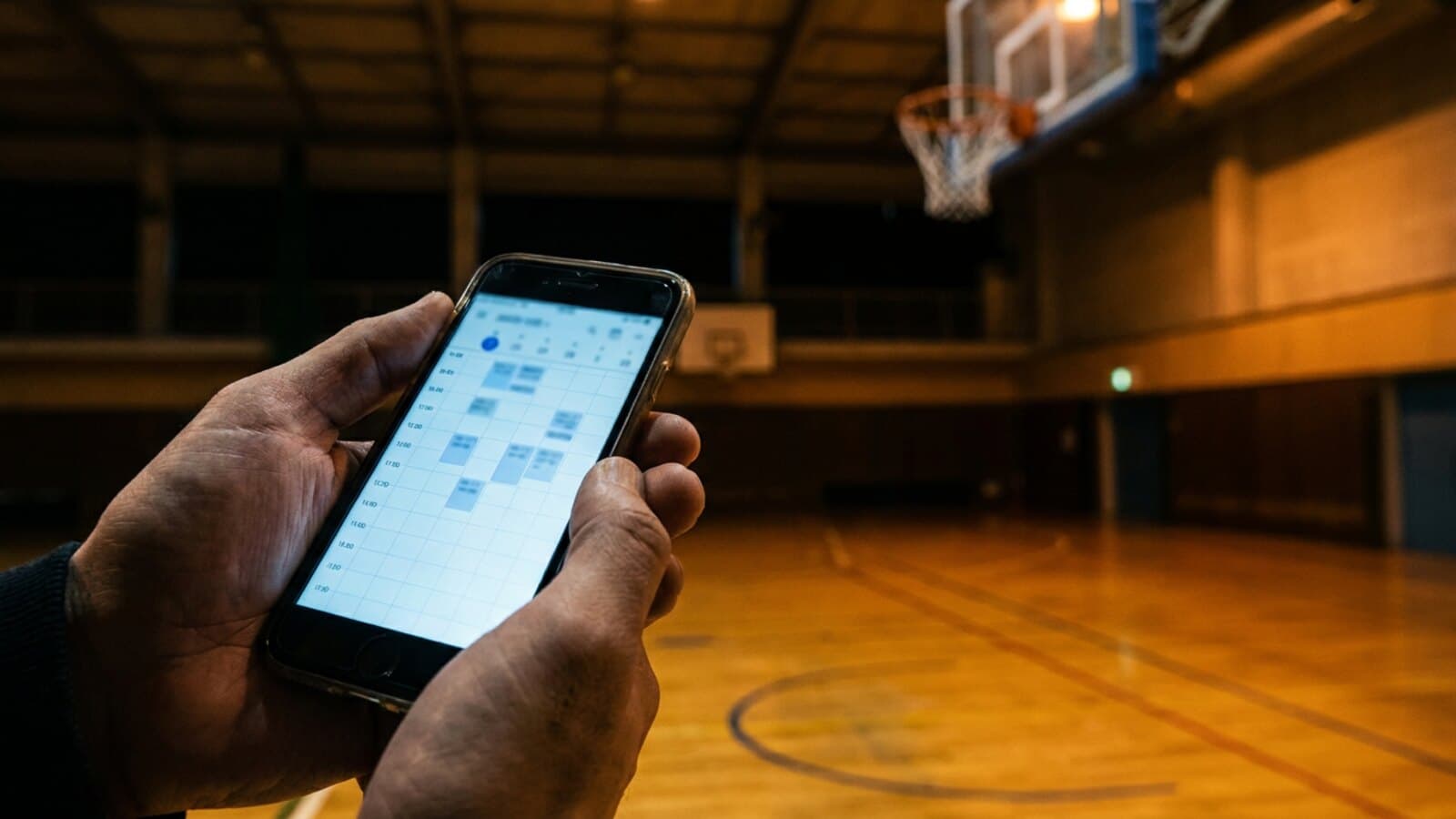 A phone held in front of an empty indoor basketball court, showing a booking screen with hour slots